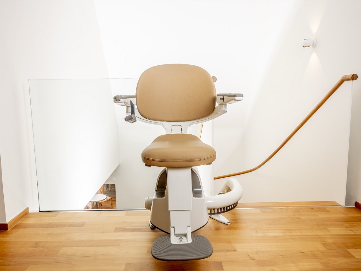 Sitzlift Flow SL mit hellem Sitz, frontal vor einer Treppe im Obergeschoss
