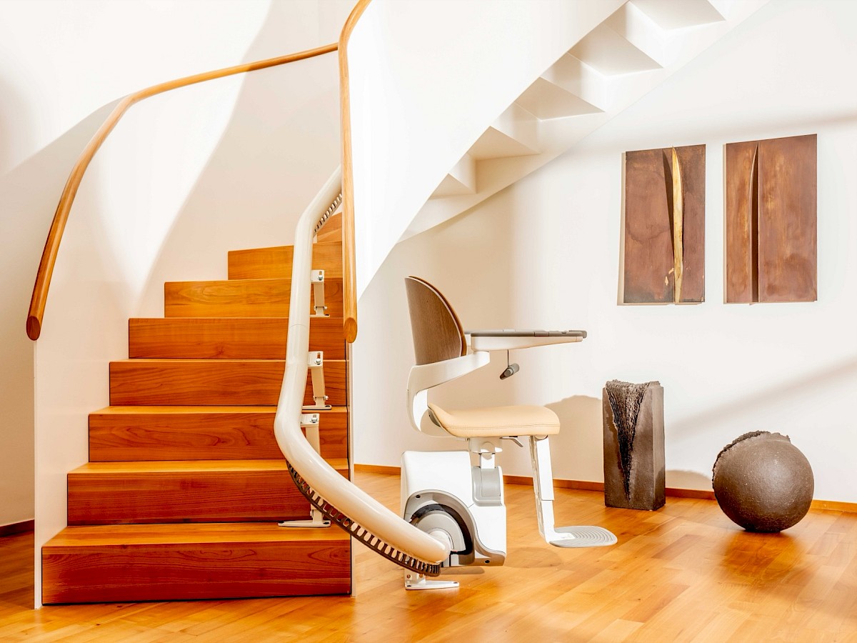 Sitzlift Flow SL mit hellem Sitz am Fu&szlig; einer geschwungenen Holztreppe in einem hellen Treppenhaus