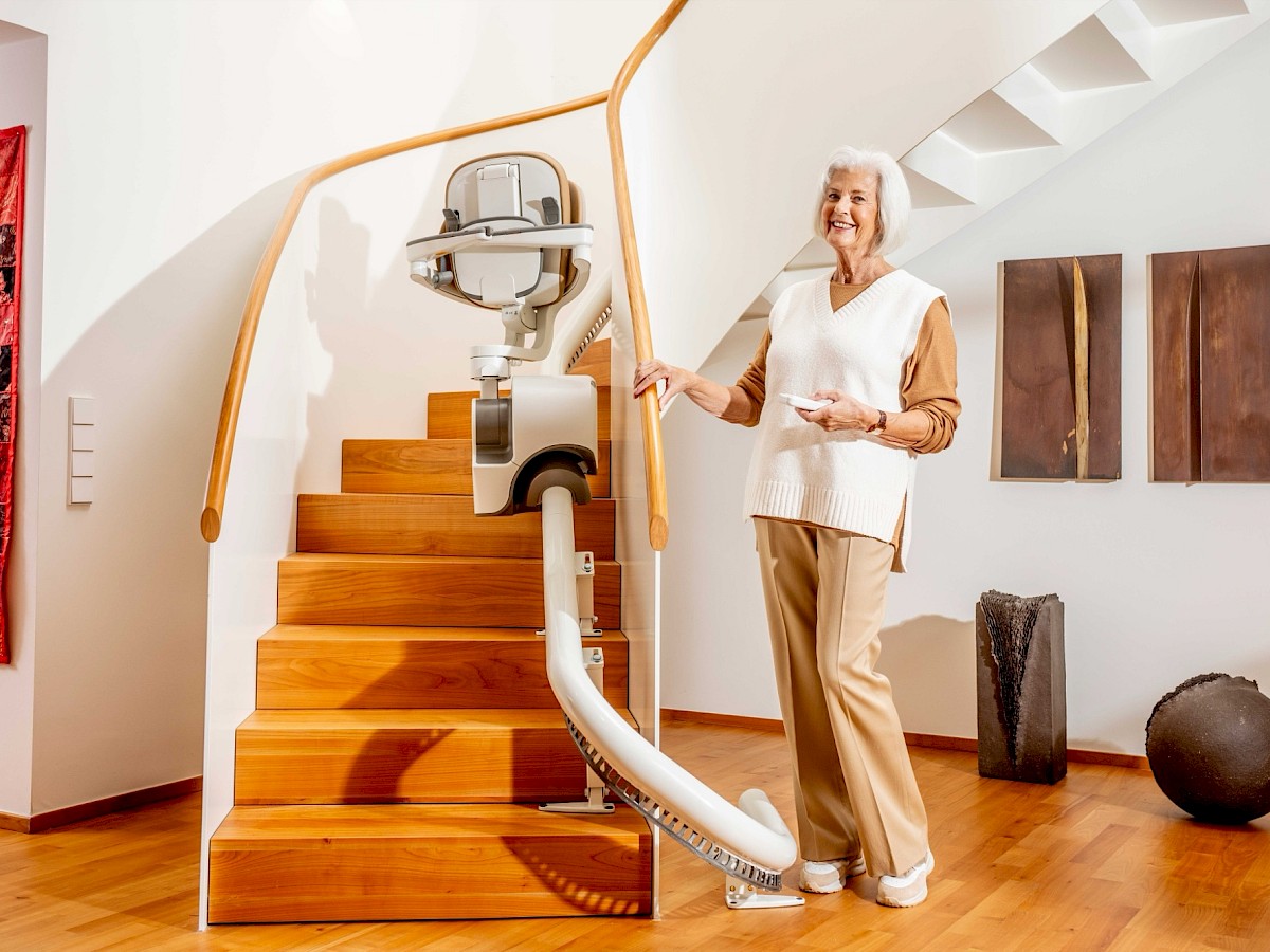 &Auml;ltere Dame steht l&auml;chelnd neben einem Sitzlift auf einer geschwungenen Holztreppe