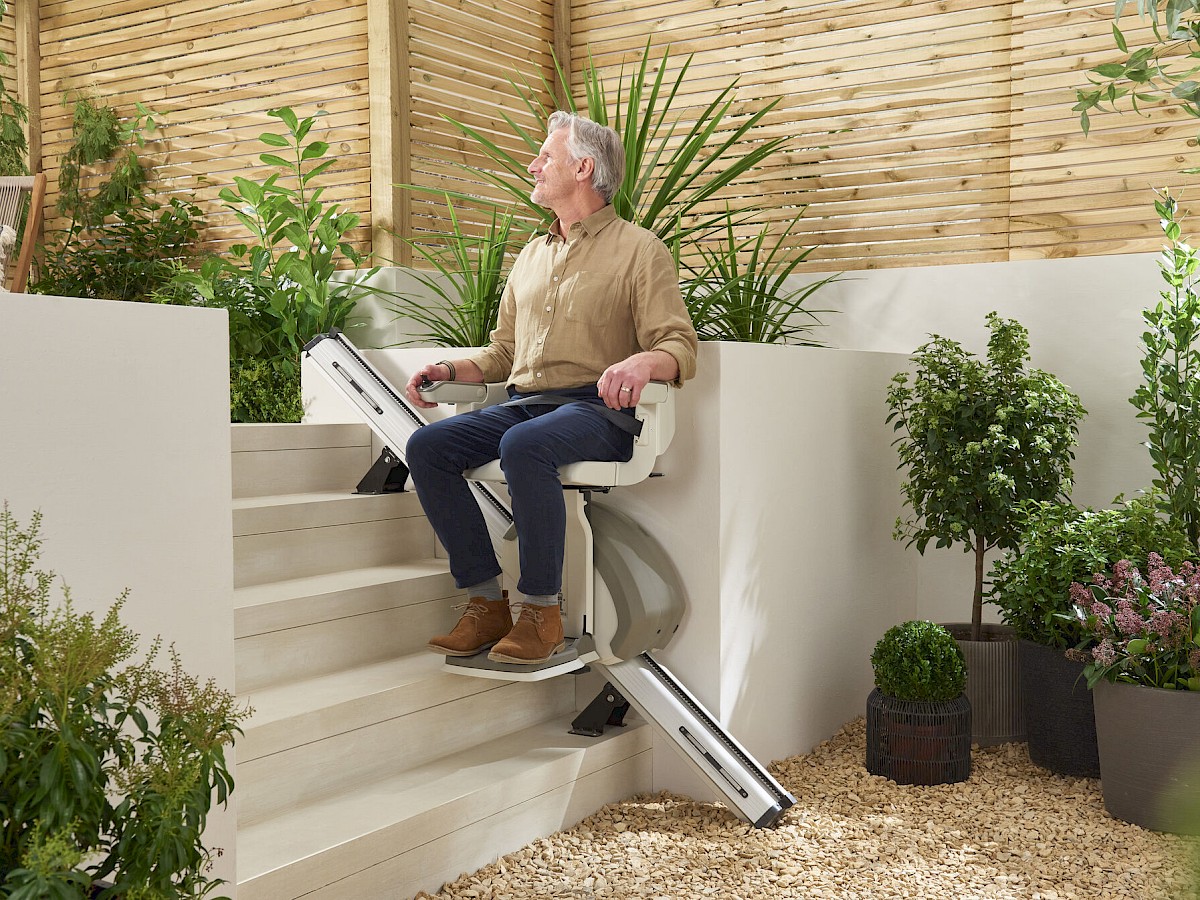Mann f&auml;hrt auf einem Sitzlift im Au&szlig;enbereich eine Treppe zwischen Pflanzen hinauf