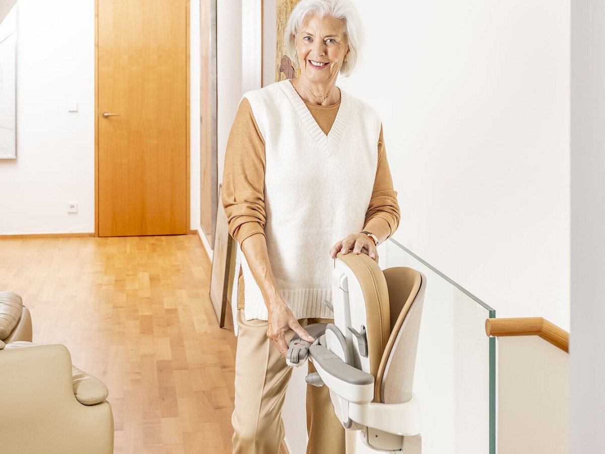 &Auml;ltere Frau l&auml;chelt neben einem Sitzlift im Flur eines Wohnhauses