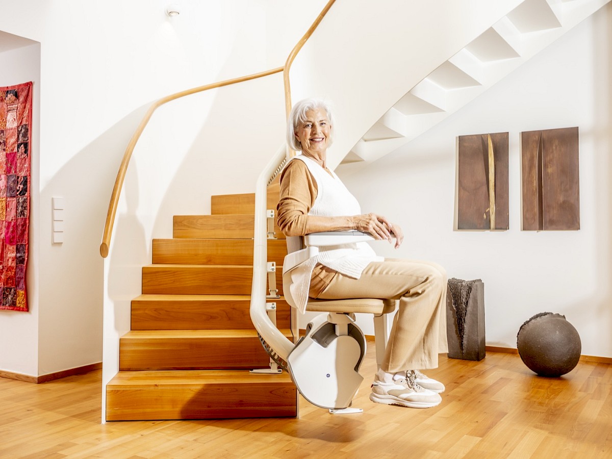 &Auml;ltere Frau sitzt auf einem Sitzlift an einer geschwungenen Holztreppe in einem hellen Treppenhaus
