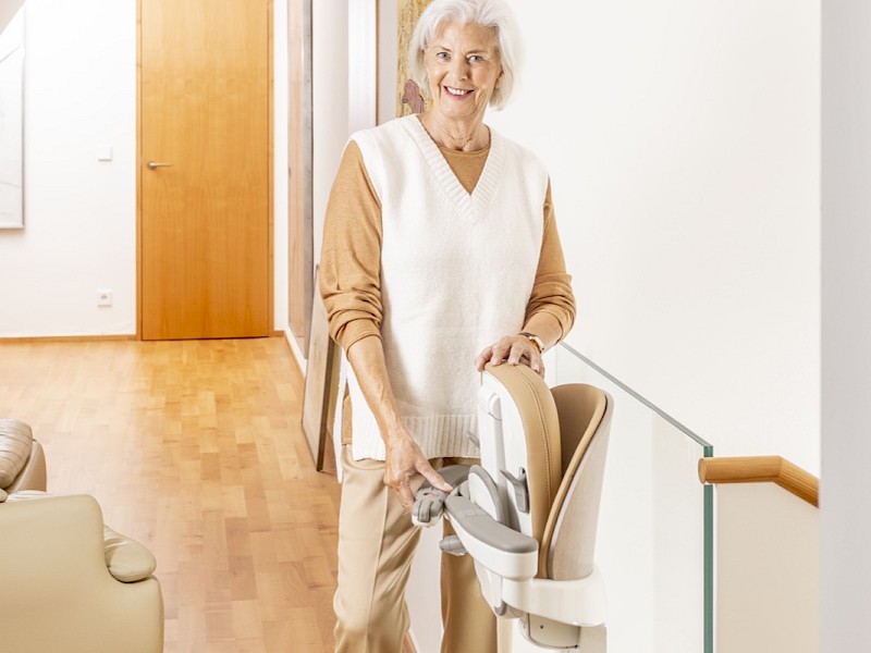 L&auml;chelnde &auml;ltere Frau steht neben einem Sitzlift im Flur eines Wohnhauses